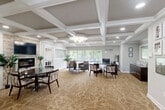 The HollySpring Ranch leasing office at 3535 Marlboro Pike in District Heights, Maryland, is a thoughtfully designed single-floor space that balances professional functionality with a warm, inviting ambiance. The interior is immediately distinguished by its striking coffered ceilings with crisp white trim, which lend an air of architectural sophistication throughout the open layout. A decorative chandelier anchors the main reception area, complemented by recessed lighting that ensures even, comfortable illumination across the space.

Patterned, neutral-toned carpeting flows throughout, adding texture and warmth underfoot. Large windows along the perimeter flood the interior with natural light and offer pleasant views of the surrounding greenery, enhancing the sense of openness. The main area features a dedicated leasing desk with guest seating, making it well-suited for resident consultations and tours.

A cozy seating lounge with upholstered armchairs provides a comfortable waiting area, while a separate dining nook with a round table and coordinating chairs is highlighted by a stone accent wall housing a wall-mounted television and fireplace, creating a welcoming focal point. A built-in kitchenette with white cabinetry and dark countertops offers added convenience. The space is completed by a welcoming entry vestibule with a glass-paneled door, framing a bright and professional first impression.