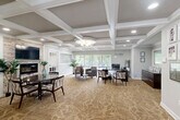 The HollySpring Ranch leasing office at 3535 Marlboro Pike in District Heights, Maryland, is a thoughtfully designed single-floor space that balances professional functionality with a warm, inviting ambiance. The interior is immediately distinguished by its striking coffered ceilings with crisp white trim, which lend an air of architectural sophistication throughout the open layout. A decorative chandelier anchors the main reception area, complemented by recessed lighting that ensures even, comfortable illumination across the space.

Patterned, neutral-toned carpeting flows throughout, adding texture and warmth underfoot. Large windows along the perimeter flood the interior with natural light and offer pleasant views of the surrounding greenery, enhancing the sense of openness. The main area features a dedicated leasing desk with guest seating, making it well-suited for resident consultations and tours.

A cozy seating lounge with upholstered armchairs provides a comfortable waiting area, while a separate dining nook with a round table and coordinating chairs is highlighted by a stone accent wall housing a wall-mounted television and fireplace, creating a welcoming focal point. A built-in kitchenette with white cabinetry and dark countertops offers added convenience. The space is completed by a welcoming entry vestibule with a glass-paneled door, framing a bright and professional first impression.