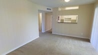 This single-floor residential space is designed with a straightforward and functional layout, making efficient use of its footprint. The living area features wall-to-wall carpeting in a neutral beige tone, creating a warm and cohesive base that complements the light-colored walls throughout. The space is finished with clean white baseboards that provide a crisp contrast to the soft, earthy palette.

The kitchen is accessible via a pass-through counter with a dark surface, which opens to the living area and promotes a sense of connectivity between spaces. White cabinetry lines the kitchen, offering ample storage, and the layout appears practical for everyday use. A ceiling-mounted light fixture provides overhead illumination in the living area.

A hallway visible from the main living space leads to additional rooms, hinting at a well-organized interior flow. The walls feature neutral beige tones throughout, contributing to a calm and understated ambiance. Ventilation is provided via ceiling-mounted HVAC registers, ensuring climate comfort year-round. Overall, the space presents a clean, move-in-ready environment with a simple, no-frills aesthetic suited to practical residential living.