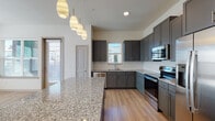This modern single-floor residence offers a thoughtfully designed open-concept layout that blends functionality with contemporary style. The kitchen serves as the heart of the home, showcasing sleek gray cabinetry, a white hexagonal tile backsplash, and polished granite countertops extending across a generous island. A full suite of stainless steel appliances — including a refrigerator with ice maker, electric range, built-in microwave, and dishwasher — completes the space. Stylish pendant lighting hangs above the island, adding warmth and character. The kitchen flows naturally into the bright living area, which features light wood-look flooring, a ceiling fan, and wide windows that invite an abundance of natural light and neighborhood views. The bedrooms are carpeted in a neutral tone, offering a comfortable retreat, with large windows providing additional natural light. The primary bathroom is a standout, featuring a dual-sink granite vanity with gray cabinetry, a large framed mirror, and a soaking tub with tile surround. A dedicated laundry area with in-unit washer and dryer hookups adds everyday convenience. Throughout, the clean white walls, consistent finishes, and light flooring create a cohesive, modern ambiance.