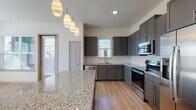 This modern single-floor residence offers a thoughtfully designed open-concept layout that blends functionality with contemporary style. The kitchen serves as the heart of the home, showcasing sleek gray cabinetry, a white hexagonal tile backsplash, and polished granite countertops extending across a generous island. A full suite of stainless steel appliances — including a refrigerator with ice maker, electric range, built-in microwave, and dishwasher — completes the space. Stylish pendant lighting hangs above the island, adding warmth and character. The kitchen flows naturally into the bright living area, which features light wood-look flooring, a ceiling fan, and wide windows that invite an abundance of natural light and neighborhood views. The bedrooms are carpeted in a neutral tone, offering a comfortable retreat, with large windows providing additional natural light. The primary bathroom is a standout, featuring a dual-sink granite vanity with gray cabinetry, a large framed mirror, and a soaking tub with tile surround. A dedicated laundry area with in-unit washer and dryer hookups adds everyday convenience. Throughout, the clean white walls, consistent finishes, and light flooring create a cohesive, modern ambiance.