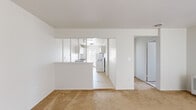 This compact yet efficiently designed one-bedroom, one-bathroom unit offers a clean, functional layout across a single floor. The combined living and kitchen area serves as the heart of the home, where a half-wall partition with a pass-through window visually connects the two spaces while maintaining a sense of definition. The galley-style kitchen features white cabinetry on both sides, gray laminate countertops with a built-in sink, white appliances including a refrigerator and electric range, and light tile flooring that brightens the space. Natural light filters in through a window at the end of the kitchen. The living area is finished with warm beige carpet, creating a comfortable and inviting atmosphere. A hallway connects the main living space to the private quarters. The bedroom is a well-proportioned room with carpeted flooring, a window with blinds for natural light, a wall-mounted air conditioning unit, baseboard heating, and a closet with louvered doors for storage. The bathroom features a wide vanity with a single basin sink, gray countertop, large wall mirror, and a shower-tub combination with a curtain. The overall aesthetic is bright and neutral throughout, with white walls and clean finishes.