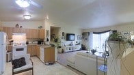 This single-floor residence showcases an open-concept layout that seamlessly connects the kitchen and living areas, creating a spacious and cohesive environment. The kitchen features warm wood cabinetry, a dark countertop peninsula that doubles as a casual dining or prep space, and white appliances including a dishwasher. A tile floor grounds the kitchen zone, transitioning into carpeted flooring in the living area.

The living space is bright and airy, benefiting from a large window that invites generous natural light and provides a view of the surrounding greenery outdoors. Neutral walls in soft beige tones complement the cream-colored sectional sofa, which anchors the sitting area around a flat-screen television mounted on a low white media console. Decorative indoor plants are distributed throughout, adding life and warmth to the space.

Recessed and flush-mount ceiling lighting supplements the natural light, ensuring the interior remains well-illuminated throughout the day. The overall ambiance is calm and functional, with an emphasis on practical living and an open, uncluttered flow between areas.