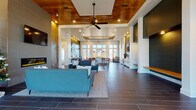 Located at 4000 Revere Blvd in Sherrills Ford, NC, this single-floor clubhouse at Revere Luxury Living offers an elegant and thoughtfully designed communal environment. The space greets visitors with a bright, double-height entry foyer featuring floor-to-ceiling windows that flood the interior with natural light, dark large-format tile flooring, and a mailbox alcove conveniently positioned near the entrance.

The open-concept main hall is anchored by a striking living area showcasing a floor-to-ceiling wood-paneled accent wall, a linear electric fireplace with a mounted TV above, and coffered ceilings with warm wood inlays and a ceiling fan. Comfortable seating arrangements with teal and charcoal sofas create an inviting lounge atmosphere throughout.

A well-appointed kitchenette area features a long white quartz island with bar seating, white cabinetry, a beverage refrigerator, and a sink — complemented by pendant lighting and a dining table with bar-height stools beneath a wood-trimmed tray ceiling. Large multi-pane windows frame exterior views throughout.

Deep green accent walls, rustic woven wall art, and warm wood tones add character and depth, while the dark hardwood-style tile flooring flows cohesively across the entire space. The overall ambiance is modern yet warm, balancing luxury finishes with approachable, community-oriented design.