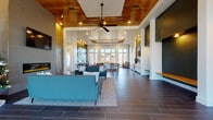 Located at 4000 Revere Blvd in Sherrills Ford, NC, this single-floor clubhouse at Revere Luxury Living offers an elegant and thoughtfully designed communal environment. The space greets visitors with a bright, double-height entry foyer featuring floor-to-ceiling windows that flood the interior with natural light, dark large-format tile flooring, and a mailbox alcove conveniently positioned near the entrance.

The open-concept main hall is anchored by a striking living area showcasing a floor-to-ceiling wood-paneled accent wall, a linear electric fireplace with a mounted TV above, and coffered ceilings with warm wood inlays and a ceiling fan. Comfortable seating arrangements with teal and charcoal sofas create an inviting lounge atmosphere throughout.

A well-appointed kitchenette area features a long white quartz island with bar seating, white cabinetry, a beverage refrigerator, and a sink — complemented by pendant lighting and a dining table with bar-height stools beneath a wood-trimmed tray ceiling. Large multi-pane windows frame exterior views throughout.

Deep green accent walls, rustic woven wall art, and warm wood tones add character and depth, while the dark hardwood-style tile flooring flows cohesively across the entire space. The overall ambiance is modern yet warm, balancing luxury finishes with approachable, community-oriented design.