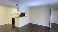 This single-floor space is designed with a clean, functional layout that emphasizes an open feel. Dark hardwood flooring runs continuously throughout, providing a rich contrast against the neutral, cream-toned walls and white trim. The living area flows seamlessly into the kitchen, which is defined by a peninsula-style breakfast bar that serves as both a workspace and a casual dining surface. The kitchen is equipped with white upper cabinetry, dark lower cabinetry, and black appliances, including a refrigerator and range, creating a two-tone aesthetic. A ceiling vent indicates a central HVAC system for climate control. Natural light enters through tall windows dressed with sheer white curtains, casting soft light across the hardwood floors. A hallway leads to additional rooms, with a doorway revealing what appears to be a closet or utility space with a darker interior finish. The overall ambiance of the space is understated and practical, with a neutral palette that offers versatility for various furnishing styles. White baseboards and door casings provide clean architectural definition throughout.