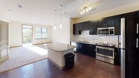 Located at 3016 Bristol Creek Dr in Morrisville, NC, this single-floor residence offers a thoughtfully designed layout with a seamless blend of style and functionality. The kitchen is a standout feature, showcasing bold dark cabinetry that extends to the ceiling, granite countertops, a subway tile backsplash, and stainless steel appliances including a refrigerator, range, and microwave. A central island with pendant lighting above serves as both a prep area and a visual anchor connecting the kitchen to the open living space. The living area is bathed in natural light through large windows, with warm carpeting underfoot and direct sightlines to the kitchen, creating an inviting and cohesive open-concept feel. Rich wood-look flooring runs through the kitchen and bathroom areas, adding warmth and continuity. The bedroom is a calm, carpeted retreat with a window that draws in natural light. The bathroom features a combination tub and shower with grab bars, dark vanity cabinetry with a light countertop, and the same wood-look flooring. A private balcony provides an outdoor retreat with views of the surrounding building courtyard, framed by metal railings under open skies.