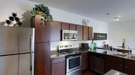 This single-story space showcases a thoughtfully designed kitchen that serves as a functional and visually appealing centerpiece. The kitchen is outfitted with rich, dark wood cabinetry that extends from floor to ceiling, maximizing storage capacity and creating a bold, warm aesthetic. The upper cabinets reach considerable heights, with decorative greenery and accents adding a lively touch to the tops of the cabinetry.

The kitchen is equipped with a full suite of stainless steel appliances, including an electric range with coil burners, an over-the-range microwave, a full-size refrigerator, and what appears to be a dishwasher. Dark granite-style countertops complement the cabinetry, providing ample workspace and a cohesive, polished look throughout. The layout follows an L-shaped configuration, offering an efficient workflow and smooth transition between cooking and prep areas.

Subtle decorative touches, including a teal glass vase and small accents, add personality to the space without overwhelming the clean design. The overall ambiance is modern and functional, with the dark tones of the cabinetry and countertops balanced by the brightness of the stainless steel finishes and light-colored walls.
