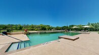This multi-story residential community is thoughtfully designed to offer both comfortable living and resort-style outdoor amenities. The property spans three floors and is anchored by a striking blue-accented clubhouse building with a steeply pitched roofline and large glass doors that open directly onto the expansive pool deck.

The centerpiece of the outdoor space is a generously sized swimming pool with a shallow entry area and a dedicated spa, all finished in natural stone coping and surrounded by a wide concrete deck. Comfortable lounge seating and large cantilever umbrellas provide shaded areas ideal for relaxing poolside. The pool area offers open views of the surrounding landscape, giving it an airy, resort-like atmosphere.

Adjacent to the pool, a covered wooden pavilion serves as an outdoor gathering hub, equipped with grilling stations and picnic-style seating — perfect for community events and social gatherings. The grounds surrounding the amenity area are well-maintained, featuring manicured lawns and open green spaces enclosed by ornamental wrought iron fencing.

Beyond the fence line, the community opens onto expansive natural terrain with mature trees and open fields, lending a sense of privacy and connection to nature. The overall setting combines functional community living with attractive outdoor spaces that encourage an active and social lifestyle.