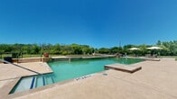 This multi-story residential community is thoughtfully designed to offer both comfortable living and resort-style outdoor amenities. The property spans three floors and is anchored by a striking blue-accented clubhouse building with a steeply pitched roofline and large glass doors that open directly onto the expansive pool deck.

The centerpiece of the outdoor space is a generously sized swimming pool with a shallow entry area and a dedicated spa, all finished in natural stone coping and surrounded by a wide concrete deck. Comfortable lounge seating and large cantilever umbrellas provide shaded areas ideal for relaxing poolside. The pool area offers open views of the surrounding landscape, giving it an airy, resort-like atmosphere.

Adjacent to the pool, a covered wooden pavilion serves as an outdoor gathering hub, equipped with grilling stations and picnic-style seating — perfect for community events and social gatherings. The grounds surrounding the amenity area are well-maintained, featuring manicured lawns and open green spaces enclosed by ornamental wrought iron fencing.

Beyond the fence line, the community opens onto expansive natural terrain with mature trees and open fields, lending a sense of privacy and connection to nature. The overall setting combines functional community living with attractive outdoor spaces that encourage an active and social lifestyle.