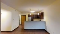 This single-floor space showcases an open-concept layout that seamlessly connects the kitchen and living area, creating a functional and inviting environment. The kitchen is outfitted with rich, dark wood cabinetry that provides ample storage, complemented by light-colored countertops that offer a striking contrast. A peninsula-style breakfast bar extends into the living area, making it ideal for casual dining or additional workspace.

Three pendant lights hang above the kitchen area — two smaller fixtures flanking a larger centered light — providing focused illumination and adding a contemporary design element. Stainless steel appliances, including a refrigerator and microwave, are integrated into the cabinetry for a streamlined appearance.

The flooring throughout is a warm hardwood-style material, lending consistency and warmth across the open space. Neutral, light-toned walls enhance the sense of openness and allow natural and artificial light to reflect evenly. A wood-toned entry door is visible in the background, framed by a short hallway. The overall aesthetic balances modern finishes with warm, natural tones.