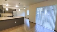 This single-floor space is designed with an open-concept layout that seamlessly connects the kitchen and living areas, creating a cohesive and functional environment. The kitchen features a dark-toned peninsula with a light countertop, providing both prep space and a natural dividing point between the cooking and living zones. A stainless steel sink and a full-size refrigerator complete the kitchen setup, while two stylish pendant lights hang above the peninsula, adding focused illumination and a modern touch. The flooring throughout is hardwood-style, lending warmth and continuity across the entire space. Natural light is a defining characteristic of this unit, with a large window and an expansive sliding glass door fitted with vertical blinds that flood the interior with brightness and offer direct access to an outdoor area. The walls are finished in a clean, neutral tone that complements the overall palette and keeps the space feeling open and airy. The layout is efficiently organized, with clear sightlines from the kitchen through to the living area, making the space feel larger than its footprint suggests. Overall, the unit offers a practical and inviting atmosphere suited for everyday living.
