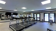 Located at 1455 Washington Blvd in Stamford, Connecticut, this fitness center offers a well-appointed, single-floor exercise facility designed to support a comprehensive workout experience. The space features a clean, modern aesthetic with a neutral color palette, dark crown molding trim, and light-toned walls that create a polished, professional atmosphere.

The cardio area is equipped with multiple treadmills and elliptical machines, each featuring individual digital display screens for tracking workout metrics. The equipment is arranged in an organized, spacious layout that allows for comfortable movement throughout the floor. A free weights section with dumbbells is also visible along the perimeter, catering to strength training needs.

Natural light filters into the space through several windows along the exterior wall, which are fitted with blinds for adjustable light control. The ceiling is outfitted with recessed, flush-panel lighting fixtures that provide consistent, bright illumination throughout the room. A wall-mounted flat-screen television adds an entertainment option for users during their workouts.

The flooring is finished with durable, cushioned gym-appropriate material, and the overall layout is thoughtfully designed to maximize usable floor space. This fitness center provides a functional and inviting environment for exercise at any level.