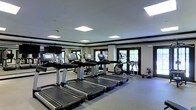 Located at 1455 Washington Blvd in Stamford, Connecticut, this fitness center offers a well-appointed, single-floor exercise facility designed to support a comprehensive workout experience. The space features a clean, modern aesthetic with a neutral color palette, dark crown molding trim, and light-toned walls that create a polished, professional atmosphere.

The cardio area is equipped with multiple treadmills and elliptical machines, each featuring individual digital display screens for tracking workout metrics. The equipment is arranged in an organized, spacious layout that allows for comfortable movement throughout the floor. A free weights section with dumbbells is also visible along the perimeter, catering to strength training needs.

Natural light filters into the space through several windows along the exterior wall, which are fitted with blinds for adjustable light control. The ceiling is outfitted with recessed, flush-panel lighting fixtures that provide consistent, bright illumination throughout the room. A wall-mounted flat-screen television adds an entertainment option for users during their workouts.

The flooring is finished with durable, cushioned gym-appropriate material, and the overall layout is thoughtfully designed to maximize usable floor space. This fitness center provides a functional and inviting environment for exercise at any level.