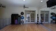 This dedicated fitness studio is thoughtfully designed to support a wide range of exercise routines in a clean, organized environment. The room features warm hardwood flooring that provides a firm yet comfortable surface ideal for workouts, stretching, and movement-based activities. A ceiling fan overhead ensures steady air circulation throughout sessions.

Natural light streams in through classic multi-pane French doors, which open to an outdoor area and lend the space an airy, welcoming feel. A large wall-mounted mirror on one side enhances the sense of openness while allowing users to monitor their form during exercise.

Equipment is neatly organized throughout the room. A vertical rack holds multiple stability balls of varying sizes, making them easily accessible, while a separate rack displays a full set of medicine balls. Rolled yoga mats and folded exercise mats are stacked in a wooden storage unit, keeping the space tidy and functional. The neutral white walls and uncluttered layout contribute to a calm, focused atmosphere well-suited for both individual training and group fitness use. Overall, this studio combines practical equipment storage with a bright, open design to create an inviting and fully functional workout environment.