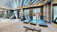 Located at 3108 McGinnis Ferry Rd in Suwanee, GA, this contemporary fitness center offers a well-appointed, single-floor workout facility designed with both function and aesthetics in mind. The space features a striking interior defined by a coffered ceiling with warm wood accents and rows of linear LED lighting that illuminate the room with a bright, energizing ambiance. Exposed concrete structural columns add an industrial character that contrasts beautifully with the polished, modern finishes throughout.

The cardio zone is anchored by a row of treadmills, elliptical trainers, and a stair climber, all positioned facing large floor-to-ceiling windows that overlook the surrounding community, allowing natural light to pour in during workouts. The strength training area is equally well-equipped, featuring Matrix cable and weight machines, a dedicated free weights section with a full dumbbell rack, adjustable benches, and a cable system with plate attachments. Floor-to-ceiling mirrors line key walls, enhancing the sense of spaciousness and allowing users to monitor their form.

The flooring transitions between patterned carpet in the strength zones and lighter surfaces near the cardio equipment, providing both comfort and practicality. Built-in cabinetry and a tiled accent wall add refined storage and design detail to one corner of the space, completing this comprehensive and attractively designed fitness facility.