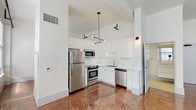 Located at 1219 E Seventh St in Los Angeles's vibrant Arts District, this single-floor unit blends contemporary design with an urban industrial aesthetic. The open living area is a standout feature, boasting polished terracotta-toned tile flooring that reflects natural light pouring in through a row of large windows. Exposed black pipe rail accents along the ceiling add an industrial touch that complements the clean white walls throughout.

The kitchen is well-appointed, featuring sleek, high-gloss white cabinetry extending toward the elevated ceiling, a marble-look stone backsplash and countertops, and a full suite of stainless steel appliances including a refrigerator, electric range, built-in microwave, and dishwasher. A modern multi-arm pendant light fixture anchors the kitchen visually, enhancing its contemporary character.

The bathroom offers a classic yet clean aesthetic, with white subway tile surrounding the soaking tub and shower combination, complemented by a charming penny-tile floor in a neutral tone. The overall ambiance of the space is bright, modern, and functional, making it well-suited for urban living in one of Los Angeles's most dynamic neighborhoods.