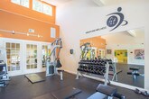 Located at 832 Blackfriars Dr in Virginia Beach, VA, this fitness and wellness center is a well-equipped, single-floor facility designed to support a comprehensive workout experience. The space is divided into distinct workout zones, each thoughtfully arranged to maximize functionality and flow. The main cardio area features multiple treadmills and an elliptical trainer lined along a wall of large sliding glass doors, allowing natural light to pour in while offering direct views of the outdoor pool area. Wall-mounted flat-screen TVs are positioned above the cardio equipment, providing entertainment during workouts. A dedicated strength training zone is outfitted with cable machines, a weight bench, a recumbent bike, and a rack of dumbbells accompanied by a wall mirror for form monitoring. An additional open area with rubber flooring provides ample space for stretching and floor exercises. The facility boasts soaring vaulted ceilings with ceiling fans, contributing to a spacious and airy atmosphere. Warm accent walls in orange and yellow, paired with vibrant, colorful artwork, give the space an energetic and inviting character. A separate adjacent room with a sink suggests restroom or changing facilities. The overall design balances practicality with an uplifting aesthetic, creating a motivating environment for residents and members alike.