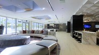 This single-floor luxury residence offers a striking open-plan layout that seamlessly integrates the living, dining, and kitchen areas into one grand, flowing space. The interior is defined by soaring ceilings adorned with layered, recessed coffers accented by integrated lighting, lending the space a refined architectural dimension. Large floor-to-ceiling windows line one side of the living area, bathing the interior in abundant natural light and offering expansive outdoor views that enhance the sense of openness.

The living area is anchored by a generous curved sectional sofa in a neutral tone, complemented by richly patterned throw pillows and a bold area rug that adds warmth and visual interest to the light hardwood flooring. Tufted ottoman seating and sleek chrome-accented coffee tables complete the sophisticated lounge setting.

The gourmet kitchen is a standout feature, showcasing dark cabinetry, open display shelving, and what appears to be a substantial island with a marble-effect surface — ideal for both culinary pursuits and casual entertaining. Dark accent walls and cabinetry in the kitchen contrast beautifully with the bright, airy palette of the surrounding living spaces. A dedicated bar or dining area with high seating is positioned to one side, adding further versatility to this exceptional residence.