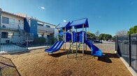 This two-story residential complex is thoughtfully designed to foster community living through a variety of shared outdoor amenities. The buildings feature a contemporary exterior with blue and gray siding accents, tile rooflines, and private balconies accessible via exterior staircases on the upper level.

At the heart of the complex lies a well-maintained communal courtyard, enclosed by a sturdy wrought iron perimeter fence with gated access points that provide both security and a welcoming sense of community. The courtyard is landscaped with lush green lawn and mature trees that offer natural shade, creating an inviting outdoor gathering area for residents.

Adjacent to the courtyard is a dedicated children's playground area, surfaced with wood chip mulch for safety. The play structure features dual blue slides, climbing elements, and covered platforms — providing an engaging space for younger residents. The playground is similarly secured within its own fenced boundary.

The complex also includes covered carport parking, ensuring convenient and protected vehicle storage for residents. Gated vehicle entry points add an additional layer of security to the property. The entire grounds benefit from abundant natural sunlight under open skies, reinforcing the bright and welcoming atmosphere of this well-appointed residential community.