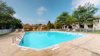 Located at 6345 Cooper Rd in Columbus, Ohio, this single-floor property features a standout outdoor amenity space centered around a generously sized swimming pool. The pool is clearly marked with depth indicators and a no-diving designation, making it suitable for recreational use by a wide range of swimmers. Surrounding the pool is a concrete deck furnished with lounge chairs, offering residents a comfortable space to relax and enjoy the outdoors. A metal fence encloses the pool area, providing both safety and a defined boundary for the amenity. Mature deciduous and evergreen trees frame the space, offering natural shade and a pleasant green backdrop under open blue skies. The surrounding residential buildings, constructed in classic brick, are visible in the background, contributing to the established, community-oriented character of the property. The interior spaces, while dimly lit in available imagery, include windowed rooms with horizontal blinds, suggesting functional residential units. Overall, the property presents a well-maintained outdoor recreational environment within a settled neighborhood setting in Columbus.