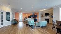 Located at 3952 Horseshoe Rd N in Little River, South Carolina, this single-floor community space combines contemporary design with a warm, welcoming ambiance. The open-concept layout seamlessly connects multiple functional zones, creating a generous and versatile gathering environment throughout.

The main lounge area is anchored by a tiled fireplace with a classic white mantel, a wall-mounted television, and vibrant abstract artwork. Comfortable seating arrangements — including teal upholstered sofas and accent chairs — are positioned around a glass-and-chrome coffee table atop a bold swirl-patterned area rug, lending the space a modern yet inviting character. Crown molding and recessed lighting add refined architectural detail throughout.

Rich hardwood-style flooring flows continuously across the entire space, tying together each zone cohesively. Natural light pours in through multiple windows and glass-paned doors, enhancing the bright, airy atmosphere. A separate seating nook with French doors provides a quieter retreat within the larger open floor plan.

The kitchen features striking navy cabinetry set against warm peach-toned walls, a spacious wraparound bar-height counter with seating, pendant lighting overhead, and black appliances. An adjacent dining area with dark wood table and upholstered chairs completes the functional gathering space, making this a well-appointed and thoughtfully designed environment for community use.