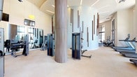 This expansive fitness center is designed with a contemporary aesthetic, combining architectural elegance with a fully equipped workout environment. Soaring ceilings and floor-to-ceiling windows along the perimeter flood the space with abundant natural light and offer views of the surrounding greenery, creating an energizing atmosphere for exercise.

The main workout floor features light-toned carpeting and is anchored by large cylindrical columns clad in wood-toned veneer. A curved accent wall adorned with vertical walnut wood dowels serves as a striking architectural focal point, complementing the clean, neutral color palette throughout. Recessed linear lighting and spherical pendant fixtures provide layered illumination across the space.

The cardio zone is well-appointed with treadmills, Matrix stair climbers, ellipticals, and stationary bikes — several positioned directly in front of large windows. The strength training area offers a wide selection of cable and selectorized machines, while a dedicated free weights room provides rubber flooring, a full dumbbell rack, a weight bench, and wall-mounted mirrors and televisions. An additional zone with rubberized flooring houses recumbent bikes and functional training equipment. Large mirrors are strategically placed throughout to enhance the sense of space and support proper form during workouts.