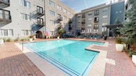 This contemporary residential community is centered around a generously sized outdoor courtyard pool, framed by a multi-story building with private balconies and arched entryways. The sparkling pool is surrounded by brick-paved decking and lined with teal lounge chairs, providing an inviting space for sunbathing and leisure. Mature trees and lush planted beds add a natural softness to the urban setting, while large potted plants and ornamental grasses enhance the landscaped borders throughout the courtyard.

An outdoor kitchen and entertainment area offers a sophisticated gathering space, featuring dual stainless steel grills, dark countertop surfaces, a bar-height counter with white stools, and built-in bench seating accented with teal and striped throw pillows. An eye-catching decorative metal lattice canopy overhead creates an artistic, semi-shaded atmosphere.

Tucked along the perimeter, a quiet garden nook features a hammock nestled beneath a mature blue spruce tree, offering a peaceful retreat. Additional landscaped beds with native plantings, white concrete benches, and brick walkways create a well-maintained and thoughtfully designed outdoor environment throughout the property.