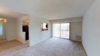 This single-floor residence offers a thoughtfully laid-out living space that balances comfort and functionality. The spacious living room is carpeted and bathed in natural light, thanks to a large sliding glass door fitted with vertical blinds that opens to an outdoor balcony area surrounded by greenery. A wall-mounted HVAC unit and ceiling vents ensure year-round climate control throughout the space.

A defining architectural feature is the pass-through window connecting the living area to the kitchen, promoting an open, communicative flow between the two spaces. The kitchen is equipped with warm wood-toned cabinetry and includes black appliances, offering a practical and welcoming cooking environment.

The neutral color palette of soft whites and beige tones carries throughout the home, creating a calm and cohesive ambiance. Smooth ceilings and clean white baseboards add a crisp, well-maintained character to the interior. The single-floor layout ensures easy accessibility and a straightforward living experience, making this space both practical and inviting for everyday living.