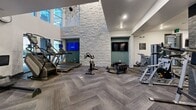 Located at 1001 Ross Ave in the heart of Dallas, TX, this fitness center offers a comprehensive workout environment within a thoughtfully designed single-floor space. The exercise room is divided into distinct functional zones, each catering to different training needs. The main workout area features an array of cardio equipment, including treadmills, stationary bikes, stair climbers, and cable machines, all arranged to maximize floor space and flow. A striking light stone accent wall serves as a dramatic architectural focal point, rising toward soaring ceiling heights in the open main section, which also features a sputnik-style chandelier and wall-mounted televisions. The flooring throughout is a herringbone-patterned carpet tile in neutral gray tones, lending a modern and polished aesthetic. Large floor-to-ceiling mirrors line the walls in multiple sections, enhancing the sense of space and aiding in form monitoring. A dedicated functional training zone offers a punching bag, kettlebells, TRX suspension trainers, a plyo box, and battle ropes. Dark stone-clad arched entryways with glass-panel double doors provide an elegant transition between zones. Recessed lighting and a clean white-and-gray color palette complete the contemporary, professional ambiance of this well-appointed fitness facility.
