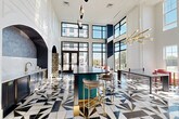 Located at 2408 South Blvd in Charlotte, NC, this single-floor space is a visually stunning communal lounge or lobby area that commands attention with its dramatic Art Deco-inspired design. Soaring double-height ceilings amplify the sense of grandeur, while floor-to-ceiling black-framed windows flood the interior with natural light and offer open views of the surrounding Charlotte cityscape.

The standout feature underfoot is a bold, geometric black, white, and gold tile floor arranged in a striking starburst and diamond pattern that anchors the entire space. Elegant arched architectural details adorn the walls, housing built-in shelving, a wet bar with marble countertops and brass fixtures, and a glass-front beverage display unit.

At the heart of the room, a teal fluted bar island with gold accents and upholstered bar stools serves as a social focal point. Seating areas are furnished with plush velvet pieces — including a mustard yellow sofa, blush pink lounge chairs, and deep red accent seating — creating an inviting, eclectic atmosphere. A large-scale floral art installation set within an arched frame adds a dramatic decorative touch. Sculptural brass sputnik chandeliers and pendant lighting complete the refined, fashion-forward aesthetic of this exceptional space.