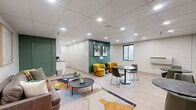 Located at 3812 Bates St in Pittsburgh's vibrant Bates Street corridor, this thoughtfully designed communal space offers a welcoming and functional environment across a single floor. The interior is defined by a striking color palette, with bold forest-green accent walls contrasting against crisp white surfaces and light wood-look flooring that runs throughout.

The space is divided into two distinct zones. A lounge area features a contemporary gray leather sofa adorned with colorful throw pillows, paired with a set of round coffee tables and anchored by a vibrant geometric area rug. Framed artwork on the white-paneled wall adds a refined, gallery-like touch. Adjacent to this is a café-style seating area furnished with dark marble-topped bistro tables and sleek green chairs, beneath a curated gallery wall of abstract paintings on the deep green accent wall.

A smaller secondary room provides a more intimate retreat, featuring a plush mustard-yellow swivel chair and a set of built-in green-painted double doors, ideal for private use or storage. Recessed lighting throughout the drop ceiling ensures bright, even illumination across all areas. A small kitchenette counter is visible along one wall, adding practical utility. Together, the spaces create a modern, inviting atmosphere well-suited for socializing, lounging, or collaborative gatherings.