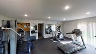 This dedicated fitness center is a single-floor space designed to support a comprehensive workout experience. The room is finished with neutral white walls and smooth ceilings fitted with recessed lighting, creating a clean and bright environment. Durable speckled rubber flooring covers the entire space, providing cushioning and traction suitable for a wide range of exercises.

The cardio section is well-stocked, featuring treadmills, an elliptical trainer, a stationary air bike, and an additional cycle machine. A multi-station cable and weight machine anchors the strength training area, complemented by a set of free weights available for targeted exercises. A bulletin board and wall clock are mounted near the entrance, adding a functional touch to the space.

Large wall-mounted mirrors span a significant portion of one wall, visually expanding the room and allowing users to monitor their form during workouts. Vertical blind-covered windows and a glass-paneled door allow natural light to filter in, offering views of the surrounding greenery outside. A separate doorway leads to an adjoining room, adding flexibility to the layout. The fitness center entrance door features a "Fitness Open" decal, clearly marking the space as a dedicated exercise facility accessible to its users.