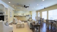 This single-floor space offers an open-concept layout that effortlessly connects the living, kitchen, and dining areas into one cohesive environment. The living area is anchored by plush, neutral-toned seating arranged around a gold-accented coffee table, creating an inviting and relaxed atmosphere. Rich hardwood flooring flows throughout, contrasting beautifully with the light, cream-colored walls that enhance the sense of openness and airiness.

The kitchenette is outfitted with white cabinetry, stainless steel appliances, and a tiled backsplash, providing a clean and functional workspace. Decorative coastal-inspired artwork adorns the walls, adding a subtle thematic character to the space. A raised counter with bar-style seating bridges the kitchen and dining areas, offering versatile options for casual meals or social gatherings.

Recessed lighting is installed throughout the ceiling, supplemented by a ceiling fan that ensures comfortable airflow. Large windows along the perimeter allow natural light to pour in, brightening every corner of the space. The overall ambiance is warm, welcoming, and well-appointed, making this an ideal multipurpose space suited for both relaxation and entertaining.