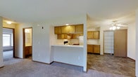 This single-floor space is designed with an open and functional layout that connects the kitchen and living areas seamlessly. The kitchen is the focal point, featuring warm honey-toned wood cabinetry that extends both above and below the counter, providing ample storage. A raised breakfast bar serves as a natural divider between the kitchen and the carpeted living area, offering a casual dining or workspace option.

The kitchen is equipped with a white electric range and refrigerator, set against light-colored walls that keep the space feeling bright and airy. Recessed lighting above the kitchen provides focused illumination. The flooring transitions from tile in the kitchen and adjacent areas to carpet in the living space, adding warmth underfoot.

Beyond the kitchen, additional cabinetry lines the wall of a secondary area, suggesting extra storage or a utility space. A closet with shelving is also visible, offering organized storage solutions. The overall color palette is neutral, dominated by whites and warm wood tones, creating a cohesive and inviting atmosphere throughout the space.