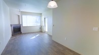 This single-floor space offers a welcoming, light-filled interior defined by clean lines and a neutral color palette. The open living area is anchored by warm hardwood-style flooring that runs throughout, lending a cohesive and contemporary feel to the space. Large windows allow generous amounts of natural light to pour in, illuminating the room and creating an airy, bright atmosphere throughout the day. A stylish pendant light fixture hangs overhead, adding a subtle design accent to the open dining and living zone. Smooth, light-toned walls provide a versatile backdrop, well-suited for a variety of interior styles. A doorway leads to an adjacent room or entryway, suggesting a functional layout that balances open communal areas with more private spaces. A wall-mounted HVAC unit ensures climate comfort year-round. The overall ambiance is calm and understated, with an emphasis on openness and natural light. The space presents itself as a clean, move-in-ready environment with thoughtful foundational details that make it adaptable to a wide range of furnishing and decorating preferences.