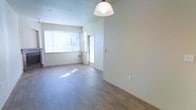 This single-floor space offers a welcoming, light-filled interior defined by clean lines and a neutral color palette. The open living area is anchored by warm hardwood-style flooring that runs throughout, lending a cohesive and contemporary feel to the space. Large windows allow generous amounts of natural light to pour in, illuminating the room and creating an airy, bright atmosphere throughout the day. A stylish pendant light fixture hangs overhead, adding a subtle design accent to the open dining and living zone. Smooth, light-toned walls provide a versatile backdrop, well-suited for a variety of interior styles. A doorway leads to an adjacent room or entryway, suggesting a functional layout that balances open communal areas with more private spaces. A wall-mounted HVAC unit ensures climate comfort year-round. The overall ambiance is calm and understated, with an emphasis on openness and natural light. The space presents itself as a clean, move-in-ready environment with thoughtful foundational details that make it adaptable to a wide range of furnishing and decorating preferences.