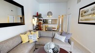 This single-floor residence showcases an open-concept layout designed for comfortable everyday living. The living area is immediately striking thanks to its soaring vaulted ceilings, which lend an expansive, airy feel to the entire space. Large windows allow abundant natural light to fill the room, highlighting the clean white walls and neutral-toned finishes throughout.

The living room provides generous space for seating and relaxation, centered around a stylish sectional sofa and a dark wood coffee table. A decorative shelving unit adds character and storage, while a pendant chandelier serves as a focal point overhead. The open layout flows naturally into the dining area, where a round table and chairs sit beneath a ceiling fan, creating an intimate yet connected space for meals.

Just beyond, the kitchen is visible through a pass-through counter, offering bar-style seating and a glimpse of the cabinetry and appliances within. The continuity between the living, dining, and kitchen areas makes the space feel cohesive and well-suited for both entertaining and daily routines. Additional doorways lead to private rooms, contributing to a thoughtful and functional floor plan throughout the residence.