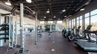 Located at 7740 Midtown Market Dr in Raleigh, NC, this fitness center offers a well-appointed, single-floor layout designed to accommodate a comprehensive range of workout needs. The facility features an open, industrial-style interior with an exposed dark ceiling structure and ductwork overhead, complemented by pendant lighting that illuminates the rubber-floored workout areas throughout.

The main workout floor is generously sized, housing a wide selection of strength training equipment, including cable machines, weight benches, and a dedicated free weights section with a full dumbbell rack. Large wall mirrors enhance the sense of space and allow users to monitor their form during training sessions. A decorative bee mural adds a distinctive visual accent to the wall.

A dedicated cardio zone lines the perimeter, featuring treadmills, ellipticals, and stationary bikes positioned along floor-to-ceiling windows that frame expansive outdoor views of the surrounding Raleigh landscape. Natural light floods these areas, creating a bright and motivating environment. Mounted televisions provide additional entertainment throughout the space.

Practical amenities include a dual-station water fountain and bottle-fill station set against sleek, large-format tile, ensuring convenience during workouts. The overall design balances functionality with a modern aesthetic, making it a well-equipped destination for fitness in the Midtown Raleigh area.