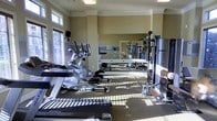 This fitness center is thoughtfully designed to support a comprehensive workout experience within a clean, well-maintained single-floor space. The room features elegant crown molding and neutral-toned walls that lend a polished, welcoming ambiance. Large windows fitted with blinds line the walls, flooding the space with generous natural light and creating an energizing atmosphere for exercise. A full wall of mirrors enhances the sense of openness and allows for proper form monitoring during workouts. The equipment selection is robust, including a Matrix treadmill, elliptical trainers, and upright cycling machines positioned along one side of the room, ideal for cardiovascular training. On the opposite side, a multi-station cable and strength training machine offers a variety of resistance exercises. The flooring is a combination of carpeting and exercise-appropriate surfaces, providing both comfort and functionality. The layout is open and organized, allowing for easy movement between equipment. Overhead lighting supplements the natural light, ensuring the space remains bright throughout the day. This fitness center is well-suited for a full-body workout routine, combining cardio, strength, and functional training capabilities in a comfortable and visually appealing environment.