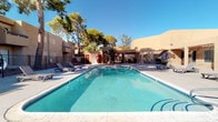 This Southwestern-inspired property showcases a striking adobe-style architectural design, characterized by warm earth-toned stucco exteriors, flat rooflines, and clean geometric forms that evoke the classic desert Southwest aesthetic. The single-story layout is thoughtfully arranged around a generous outdoor communal area that serves as the centerpiece of the space.

The outdoor area features a large, crystal-clear swimming pool with vivid turquoise water, framed by a wide surrounding deck finished in light-toned pavers. Lounge chairs are positioned poolside, offering ample space for relaxation under the open sky. A separate spa is also visible adjacent to the main pool. Tall, mature palm trees and established shade trees rise above the roofline, adding natural texture and a resort-like ambiance to the setting.

The building facades bordering the pool area incorporate covered pergola-style overhangs, large windows, and decorative tile accents, blending functional architecture with visual character. Gated fencing encloses the pool area, ensuring a secure and private environment. Blue canvas umbrellas provide shaded gathering spots on the deck.

Abundant natural sunlight floods the outdoor space throughout the day, reflecting brilliantly off the pool surface and highlighting the warm tones of the surrounding architecture. The overall atmosphere is open, bright, and inviting, with a strong sense of desert charm.