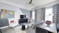 This single-floor residence showcases a thoughtfully designed living area that blends modern aesthetics with comfortable functionality. The focal point of the living room is a striking geometric accent wall rendered in cool grey and white tones, creating a bold yet sophisticated visual statement. A sleek, dark wood media console anchors the space, providing both storage and a platform for the flat-screen television.

The room is furnished with a generous sectional sofa in a soft grey upholstery, complemented by a crisp white accent chair adorned with red patterned cushions, introducing pops of color that energize the neutral palette. A textured area rug grounds the seating arrangement, while a round metal side table adds an industrial-modern touch.

Natural light filters in through multiple windows dressed in classic plaid drapery, lending warmth and pattern to the space. A modern ceiling fan with a brushed metallic finish provides both airflow and a refined design detail overhead. The light hardwood-style flooring ties the entire space together, enhancing the bright and airy ambiance throughout the room.