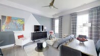 This single-floor residence showcases a thoughtfully designed living area that blends modern aesthetics with comfortable functionality. The focal point of the living room is a striking geometric accent wall rendered in cool grey and white tones, creating a bold yet sophisticated visual statement. A sleek, dark wood media console anchors the space, providing both storage and a platform for the flat-screen television.

The room is furnished with a generous sectional sofa in a soft grey upholstery, complemented by a crisp white accent chair adorned with red patterned cushions, introducing pops of color that energize the neutral palette. A textured area rug grounds the seating arrangement, while a round metal side table adds an industrial-modern touch.

Natural light filters in through multiple windows dressed in classic plaid drapery, lending warmth and pattern to the space. A modern ceiling fan with a brushed metallic finish provides both airflow and a refined design detail overhead. The light hardwood-style flooring ties the entire space together, enhancing the bright and airy ambiance throughout the room.