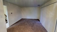 This two-story space presents a straightforward and functional design throughout its layout. The pictured room showcases wall-to-wall carpeting in a warm, neutral tan tone, providing a soft underfoot feel that contributes to a quiet, comfortable atmosphere. The walls are finished in a clean, crisp white, offering a blank canvas that suits a wide range of interior styles and furnishing choices. A ceiling-mounted HVAC vent is present, ensuring climate control throughout the space. Vertical blinds on what appears to be a sliding door or large window offer adjustable light control and privacy. The room is generously proportioned, providing ample space for a variety of furniture arrangements. The dark baseboard trim offers a subtle contrast against the lighter walls and carpet, adding a modest architectural detail. The two-floor configuration of the overall property suggests additional living or functional areas beyond what is visible in this image, allowing for separation of living spaces across levels. The overall aesthetic is neutral and move-in ready, offering flexibility for residents to customize the space to their preferences.