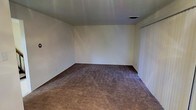 This two-story space presents a straightforward and functional design throughout its layout. The pictured room showcases wall-to-wall carpeting in a warm, neutral tan tone, providing a soft underfoot feel that contributes to a quiet, comfortable atmosphere. The walls are finished in a clean, crisp white, offering a blank canvas that suits a wide range of interior styles and furnishing choices. A ceiling-mounted HVAC vent is present, ensuring climate control throughout the space. Vertical blinds on what appears to be a sliding door or large window offer adjustable light control and privacy. The room is generously proportioned, providing ample space for a variety of furniture arrangements. The dark baseboard trim offers a subtle contrast against the lighter walls and carpet, adding a modest architectural detail. The two-floor configuration of the overall property suggests additional living or functional areas beyond what is visible in this image, allowing for separation of living spaces across levels. The overall aesthetic is neutral and move-in ready, offering flexibility for residents to customize the space to their preferences.