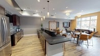 This well-appointed single-floor community amenity space is designed to provide residents with a comfortable and functional gathering environment. The open-concept layout seamlessly connects a fully equipped kitchen, a casual dining area, and a relaxed lounge zone, all unified by warm wood-look flooring throughout.

The kitchen area is outfitted with dark espresso cabinetry, sleek black granite countertops, a mosaic tile backsplash, stainless steel appliances including a full-size refrigerator and built-in microwaves, and a spacious island with pendant lighting overhead. A striking linear fireplace with floor-to-ceiling tile surround serves as a focal point along one wall, adding warmth and visual interest.

The lounge area is furnished with a tufted leather sofa, upholstered armchairs, and a rustic wood coffee table, arranged around a wall-mounted flat-screen television. Large windows flood the seating area with natural light and provide views of an adjacent outdoor patio and surrounding buildings.

The dining zone features a mix of tables and chairs in warm amber and neutral tones, complemented by a decorative wall-mounted game board. Recessed lighting and statement pendant fixtures provide layered illumination throughout the space, contributing to an inviting and modern ambiance suited for socializing, dining, or relaxing.