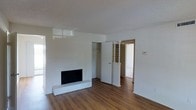This single-floor space presents a clean, minimalist aesthetic defined by crisp white walls and warm hardwood flooring that runs throughout. The main living area is anchored by a classic fireplace set into a smooth white accent wall, adding character and a focal point to the room. A built-in closet with a standard hanging rod provides practical storage directly off the living space, while a decorative lattice-style room divider adds a subtle architectural detail that helps define the transition between areas. The open doorway beyond leads to an adjacent room, suggesting a functional layout with a natural flow between spaces. The white walls and light-toned ceiling work together to create a bright, airy atmosphere, while the smooth wall surfaces lend the space a polished, well-maintained appearance. The overall ambiance is understated and versatile, offering a neutral canvas that can accommodate a variety of uses and interior styles.