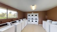 This shared laundry facility is a practical, single-floor space designed to accommodate multiple users simultaneously. The room is finished with warm brown walls and light-toned vinyl tile flooring, creating a clean and functional environment. Overhead fluorescent lighting ensures the space is well-lit at all times, complemented by natural light streaming in through several large windows that offer pleasant outdoor views of surrounding greenery.

The facility is outfitted with a generous row of top-loading washing machines lining both sides of the room, providing ample capacity for high-volume laundry needs. At the far end, three sets of stacked commercial-grade dryers are neatly arranged against the wall, maximizing the use of vertical space. Each machine features coin-operated controls, making the facility straightforward and easy to use.

A folding counter or table area is available along one wall, offering a convenient surface for sorting and folding laundry. The open floor plan allows for comfortable movement between machines, and the room maintains a tidy, organized layout throughout. A utility door provides additional access, adding to the overall functionality of the space. This laundry facility is well-suited for a residential community setting, combining efficiency, capacity, and convenience in a single, accessible room.