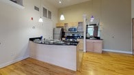 Located at 921 Commerce St in Petersburg, Virginia, this single-floor unit showcases a compelling blend of industrial heritage and contemporary design. Exposed red brick walls serve as a defining architectural feature throughout the space, complementing the warm, light-toned hardwood floors that extend across the main living and sleeping areas. The open layout creates a generous, airy feel, enhanced by a ceiling fan and natural light streaming through large windows fitted with blinds.

The open-concept kitchen is a functional centerpiece, featuring light maple cabinetry, dark granite countertops, stainless steel appliances including a refrigerator, microwave, and range, and a curved kitchen island with a built-in sink. Pendant lighting hangs above the island, adding a modern touch, while decorative columns and glass shelving provide additional architectural detail.

The bathroom is well-appointed with tile flooring, a wood-toned vanity topped with dark granite, a large mirror, and a glass-enclosed shower. Throughout the unit, the interplay of raw brick, crisp white walls, and warm wood flooring creates a loft-inspired ambiance that balances historic character with modern comfort.