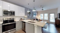 This single-floor space showcases a thoughtfully designed open-concept layout that integrates the kitchen and living area into one cohesive environment. The kitchen is a standout feature, equipped with stainless steel appliances including a built-in microwave, dishwasher, and range. Crisp white cabinetry with a high-gloss finish lines the walls, complemented by clean white quartz countertops that extend into a peninsula with an undermount sink — ideal for both meal preparation and casual dining. Two pendant lights in a muted sage tone hang above the peninsula, adding a stylish focal point while providing functional task lighting. The grey accent wall serves as a visual anchor, lending depth and contrast to the otherwise bright, neutral palette. Wood-look flooring runs throughout, adding warmth and continuity across the space. The living area beyond benefits from natural light streaming through large windows with exterior views, creating an airy, open ambiance. A ceiling fan is visible in the living zone, enhancing comfort. Overall, the space presents a clean, contemporary aesthetic with functional, well-appointed details throughout.